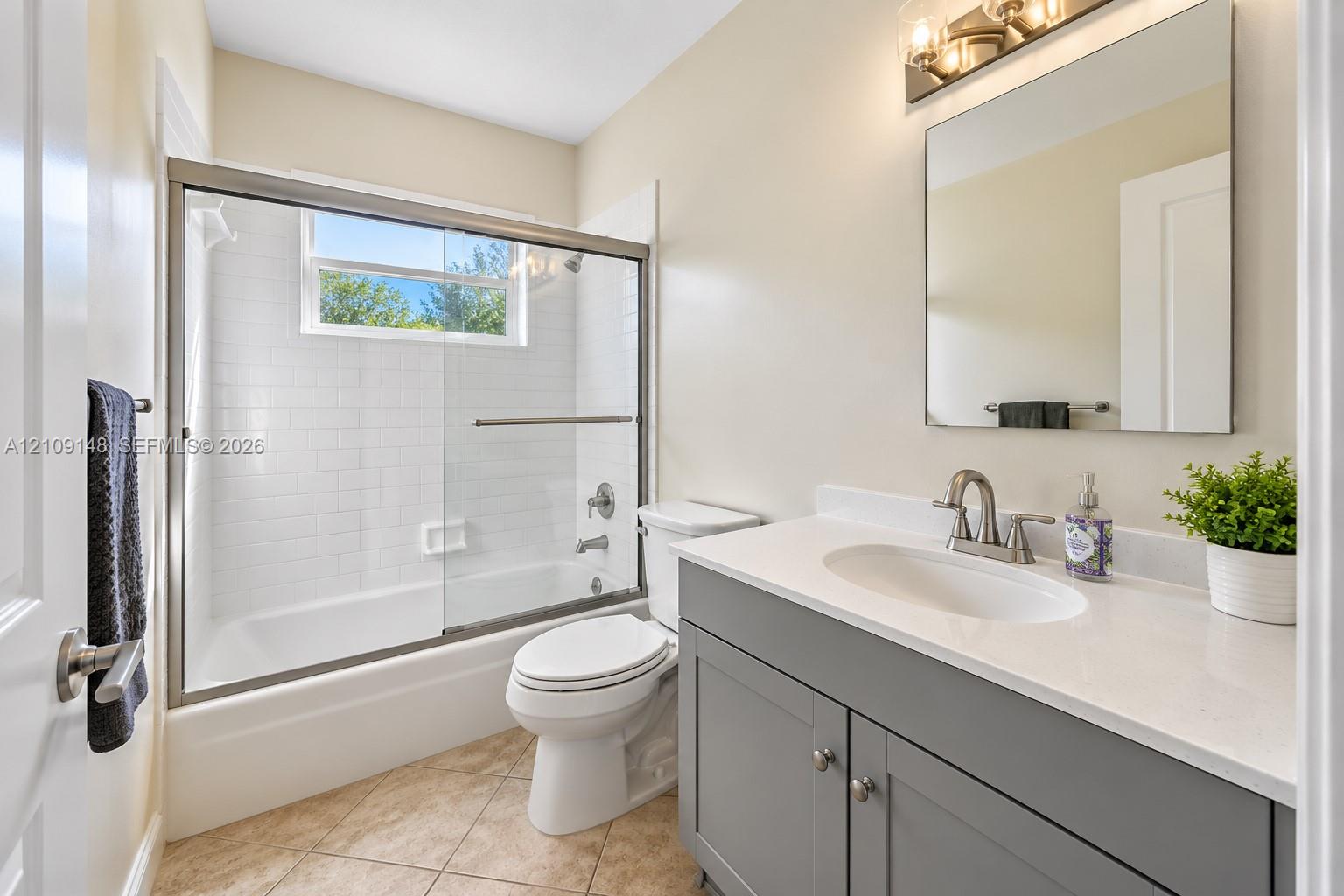 Bathroom featuring a gray vanity with a neutral solid-surface countertop, an undermount oval sink, a frameless mirror with mounted lighting, and a bathtub with a white subway-tile surround and a sliding glass enclosure