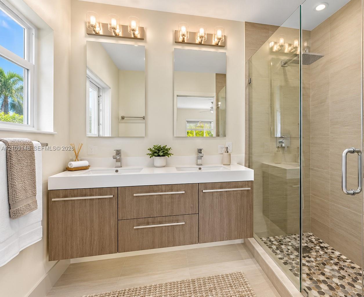 Dual-sink vanity with wood-finish cabinetry, quartz countertop, framed glass shower enclosure, stone pebble shower flooring, and wall-mounted vanity lighting