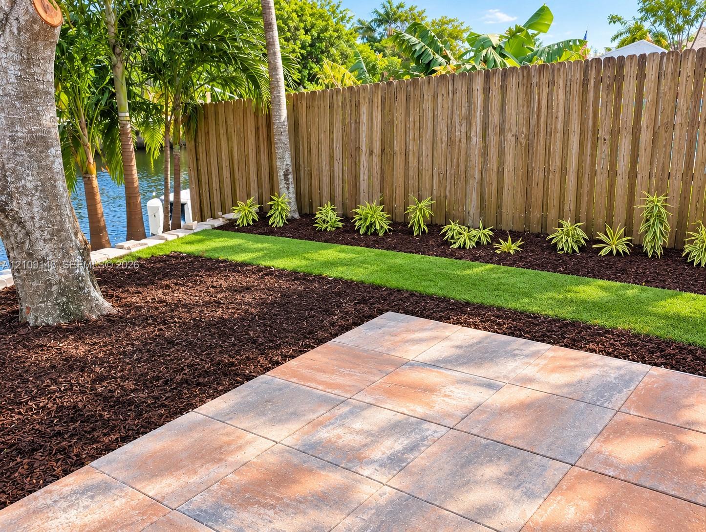 Backyard featuring large-format paver patio, manicured lawn, mulched garden beds, and perimeter wood fencing