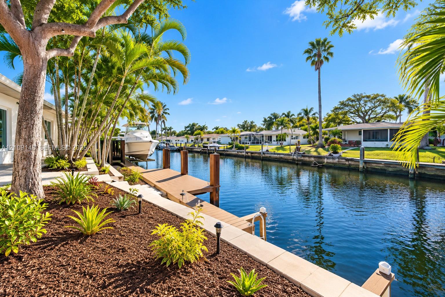 Waterfront property featuring a private wooden dock, concrete seawall, tiered landscaping with mulch beds, and mature palm trees