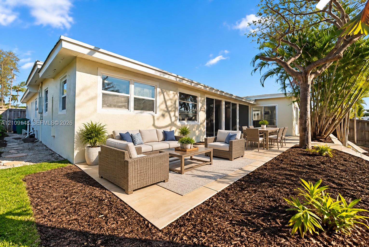 Rear exterior featuring a stucco facade, expansive paved patio, mature trees, and sliding glass door access
