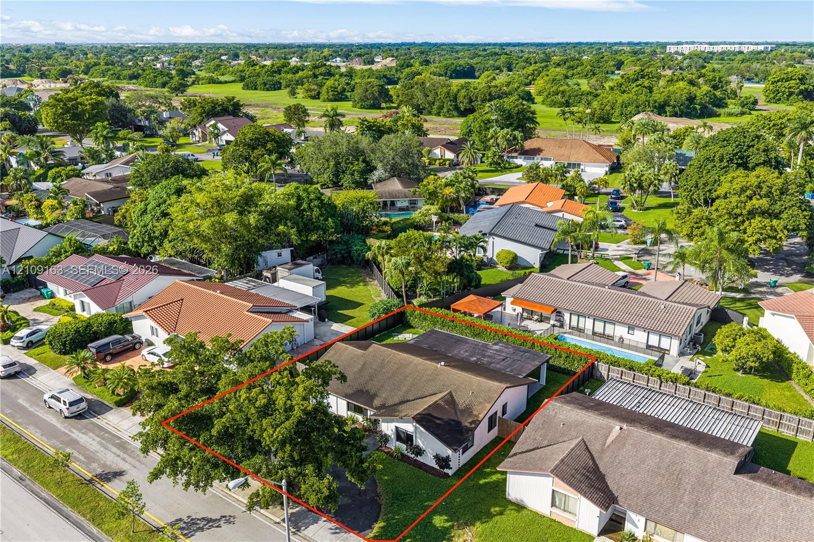 Aerial perspective of a property featuring a single-story residence, asphalt driveway, manicured lawn, and mature trees enclosed by perimeter hedging