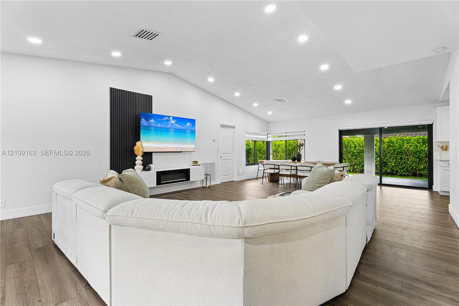 Open-concept interior featuring vaulted ceilings, wood-finish flooring, recessed lighting, a built-in electric fireplace with vertical slat wall detailing, and sliding glass doors
