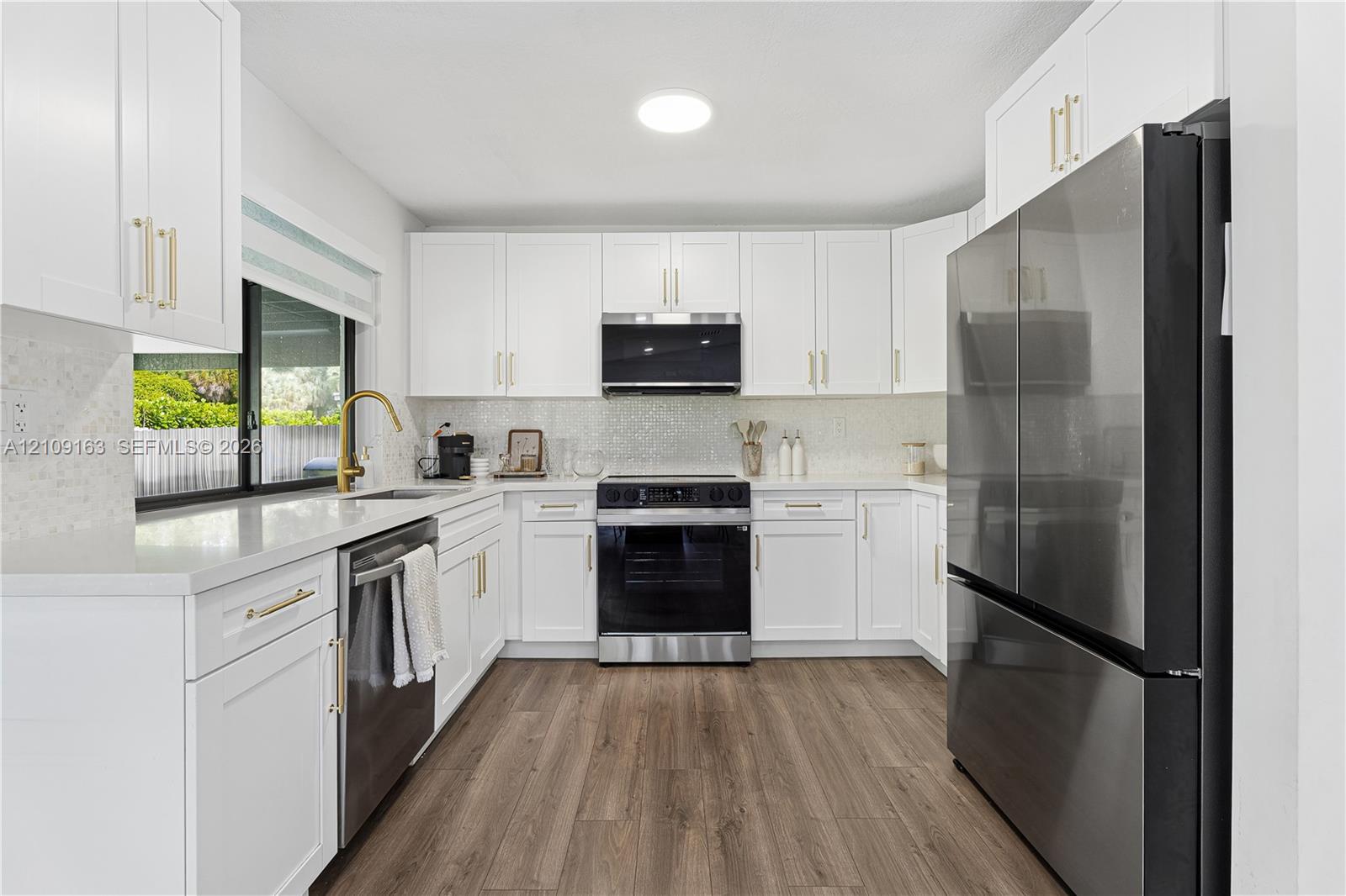 Kitchen featuring shaker-style white cabinetry, quartz countertops, stainless steel appliances, brass-finish hardware, and wood-finish flooring