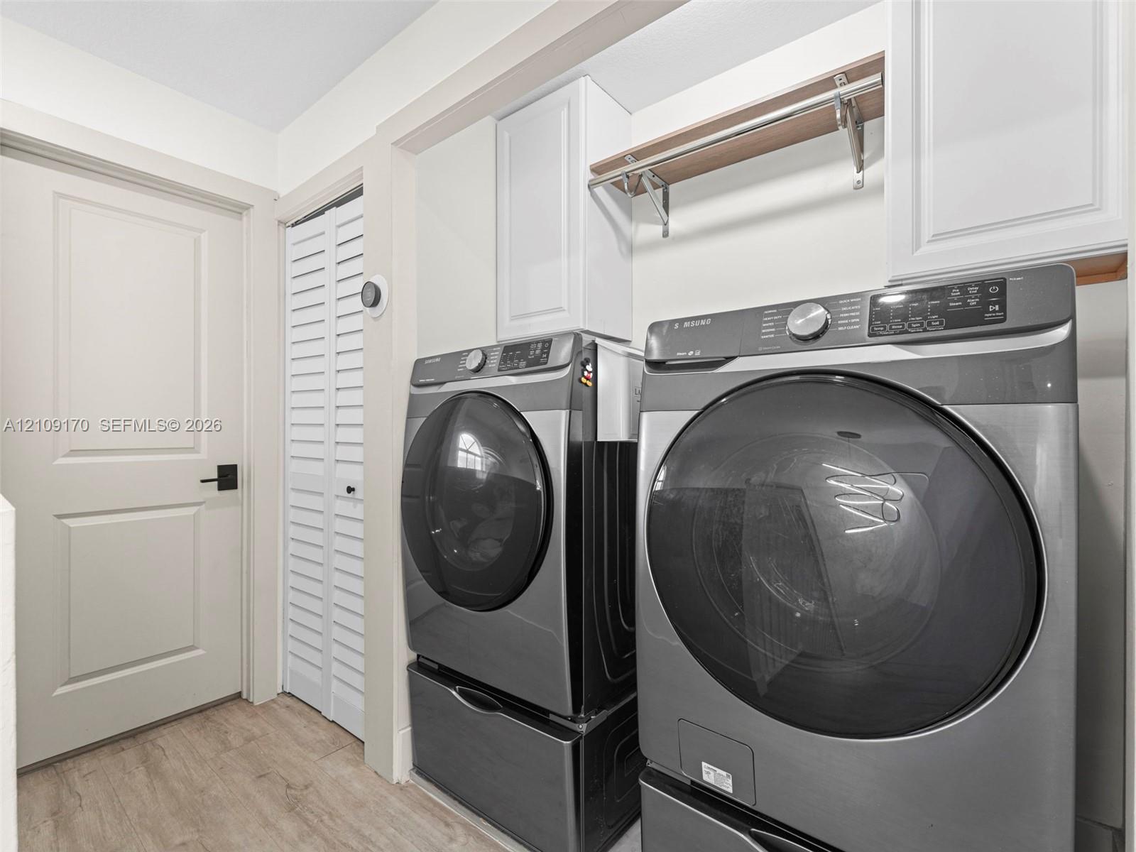Laundry space featuring white shaker-style cabinetry, a built-in wall-mounted shelving unit with integrated hanging rod, louvered closet doors, and wood-finish flooring