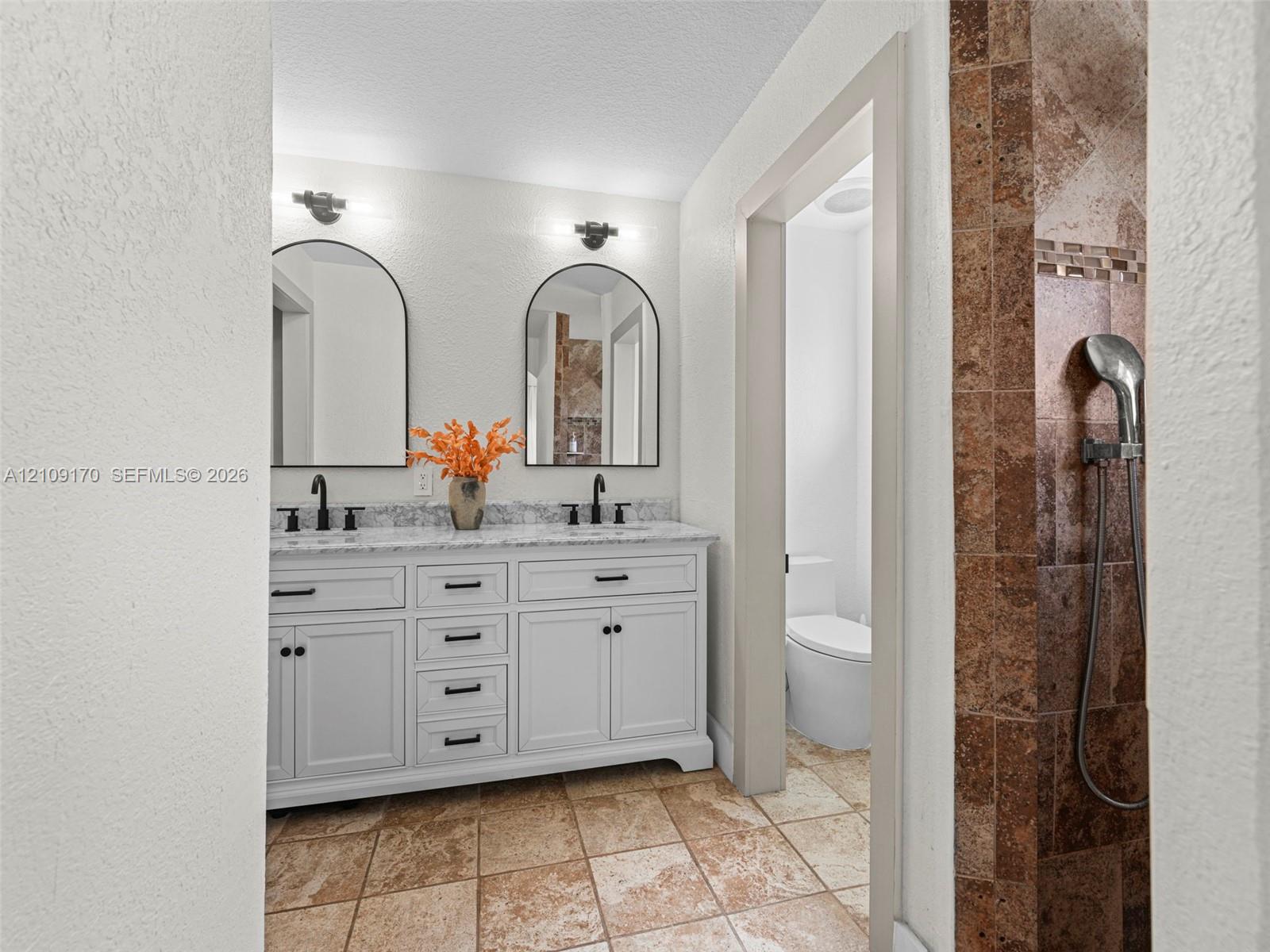 Double vanity featuring white cabinetry, stone-patterned countertop, dual undermount sinks, and matte black hardware, accented by arched mirrors and wall-mounted light fixtures, with stone-tiled flooring and a walk-in shower
