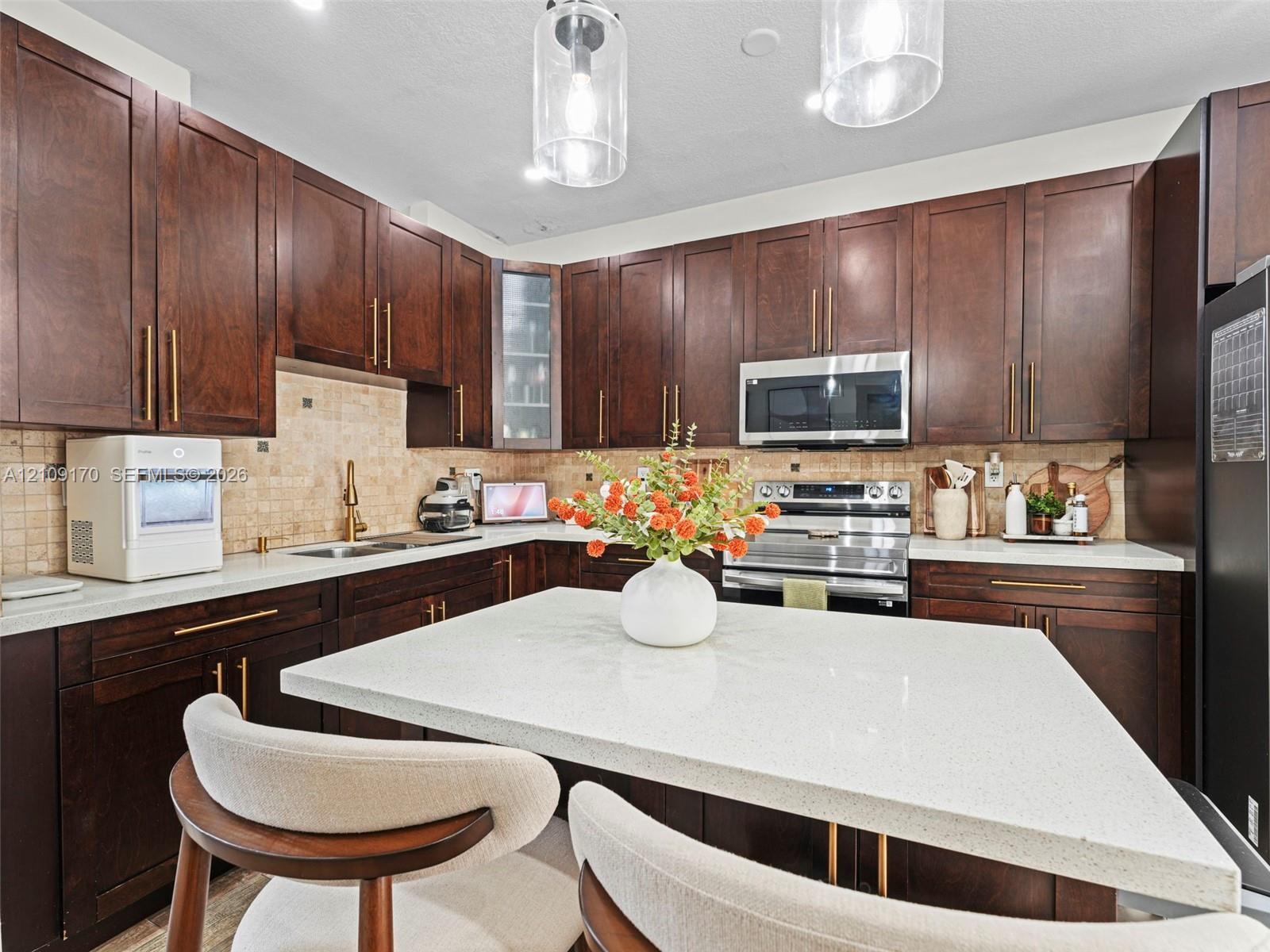 Kitchen featuring dark wood cabinetry, light quartz countertops, stainless steel appliances, a tiled backsplash, and glass pendant lighting