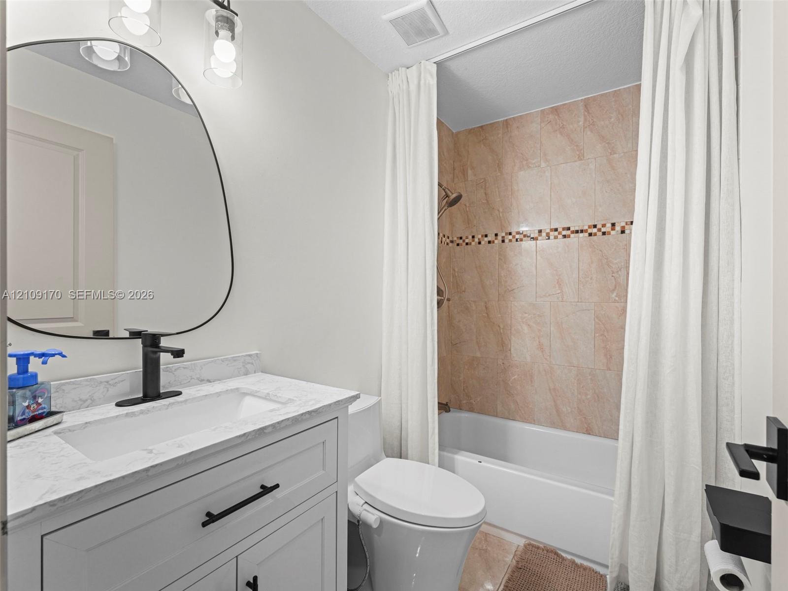 Vanity featuring a stone-finish countertop, undermount sink, and matte black faucet, paired with an asymmetrical wall mirror and tiled shower surround with mosaic accent detailing