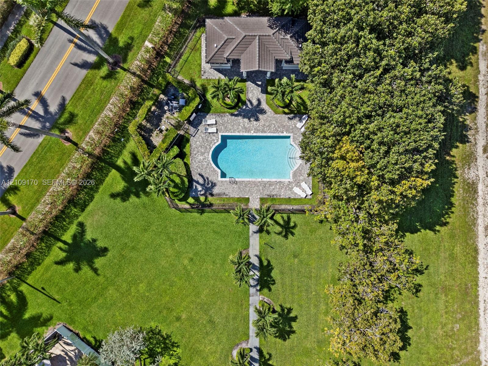 Aerial perspective of a property featuring an inground swimming pool, stone-paver pool deck, concrete walkways, mature trees, and expansive lawn area