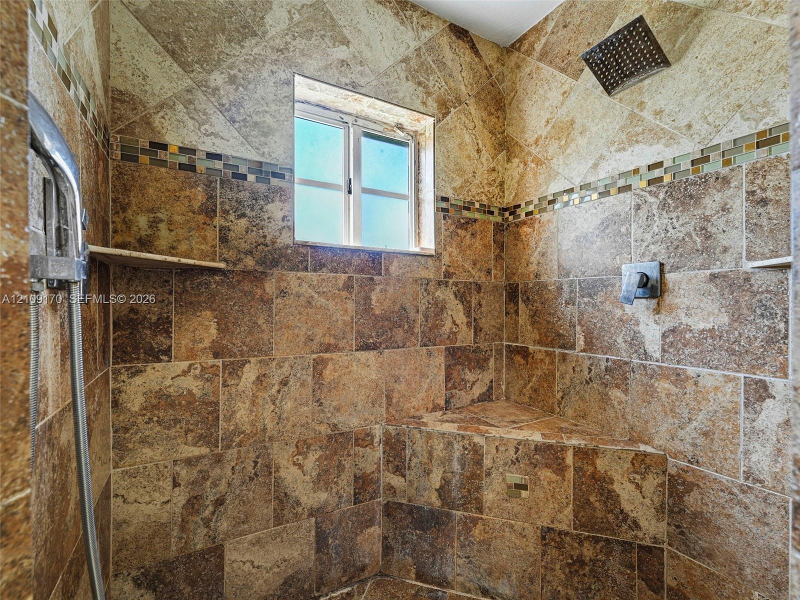 Walk-in shower featuring natural stone wall tile, an integrated corner seat, a rain-style showerhead, a handheld shower fixture, and decorative glass mosaic tile accents