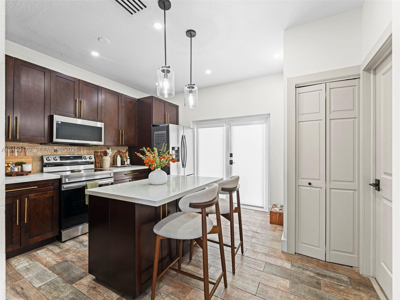 Kitchen featuring dark wood cabinetry, a central island with a stone-surface countertop, stainless steel appliances, dual glass pendant lights, and wood-finish tile flooring