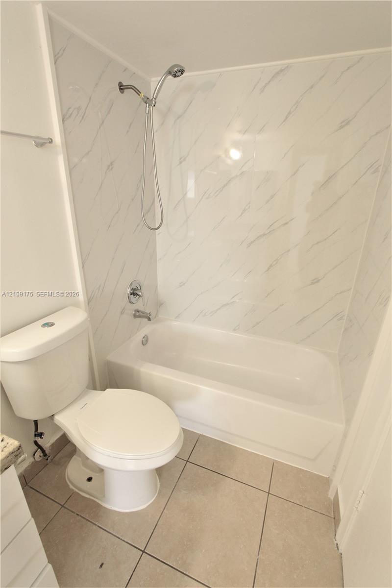 Full-size bathtub with white wall surround featuring veined detailing, handheld shower fixture, standard toilet, and neutral-toned floor tiling