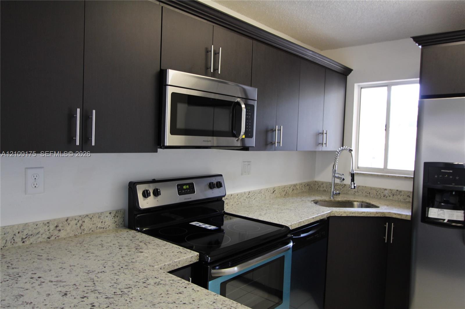 Kitchen featuring dark-toned cabinetry, light stone-patterned countertops, stainless steel appliances, a corner-mounted sink with a pull-down faucet, and crown molding