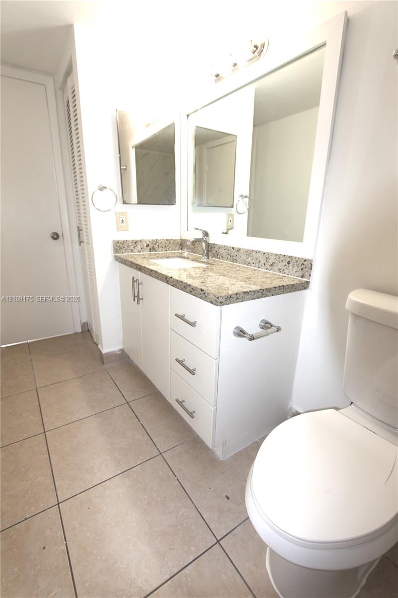 Bathroom featuring a white vanity cabinet with drawers and lower storage, stone-finish countertop, undermount sink, wall-mounted mirror with integrated lighting, and neutral-tone tile flooring