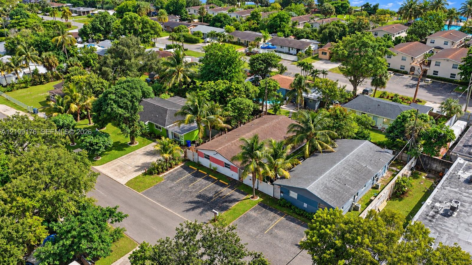 Aerial perspective of the property featuring a paved off-street parking area, multi-tone exterior siding, and a shingled roof, surrounded by mature palm trees and established landscaping