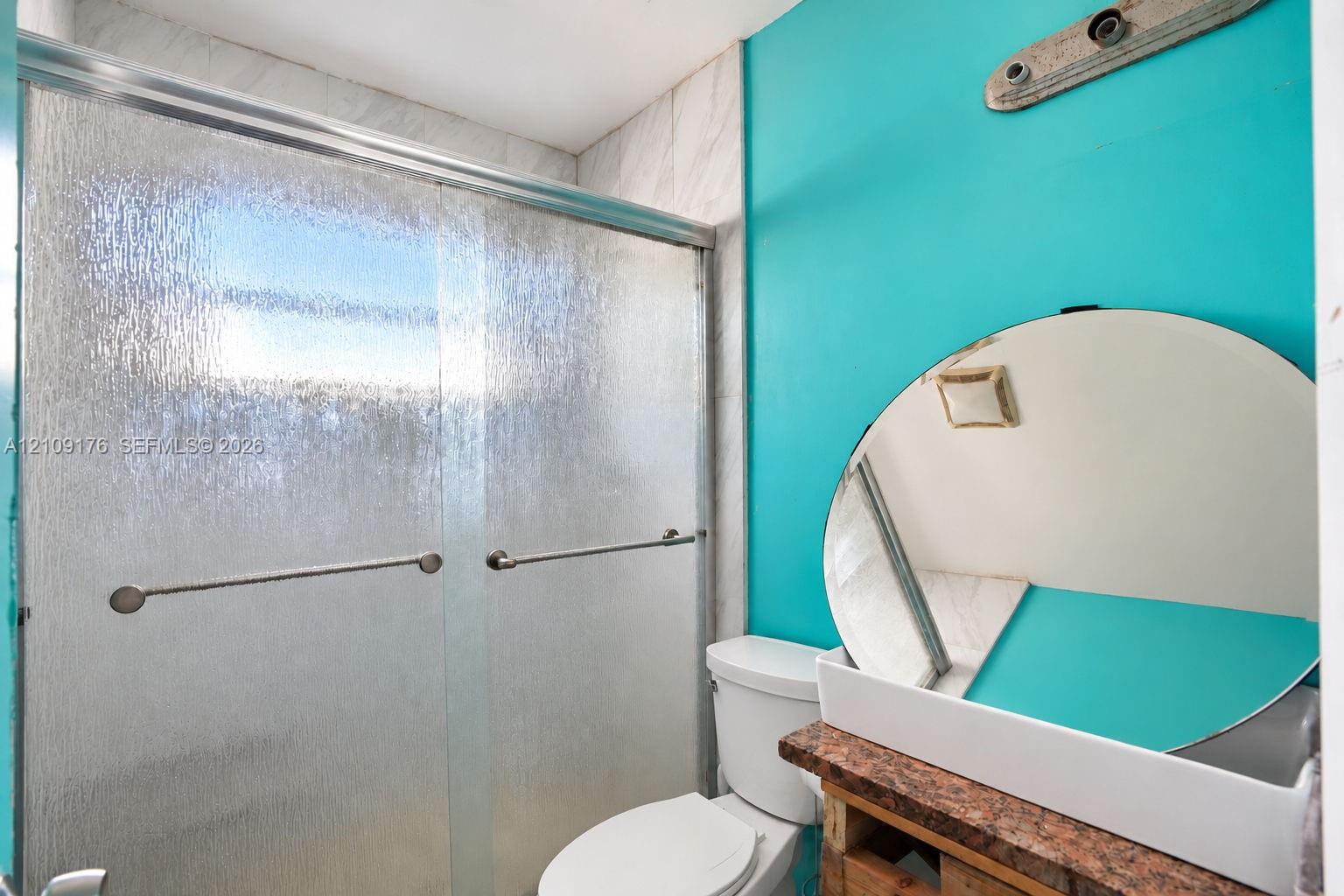 Bathroom featuring a glass shower enclosure with textured panels, a circular wall mirror, and a stone-surface vanity countertop