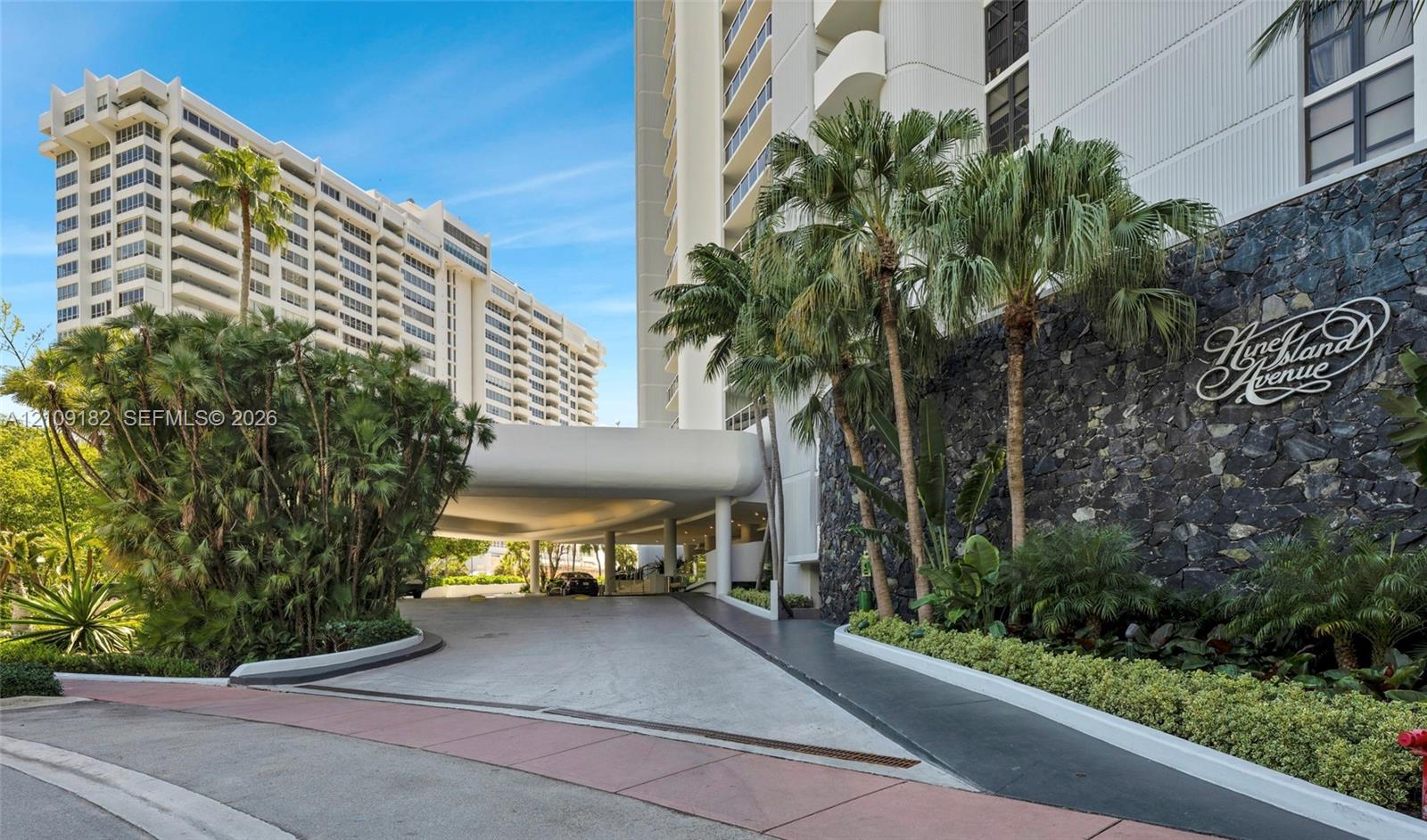 Multi-story high-rise exterior featuring a concrete porte-cochre, decorative stone masonry wall, mature palm trees, and landscaped perimeter hedging