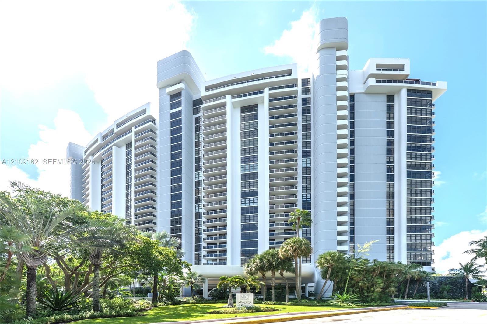 High-rise architectural exterior featuring tiered balconies, floor-to-ceiling glass windows, concrete structural columns, and landscaped grounds with mature palm trees