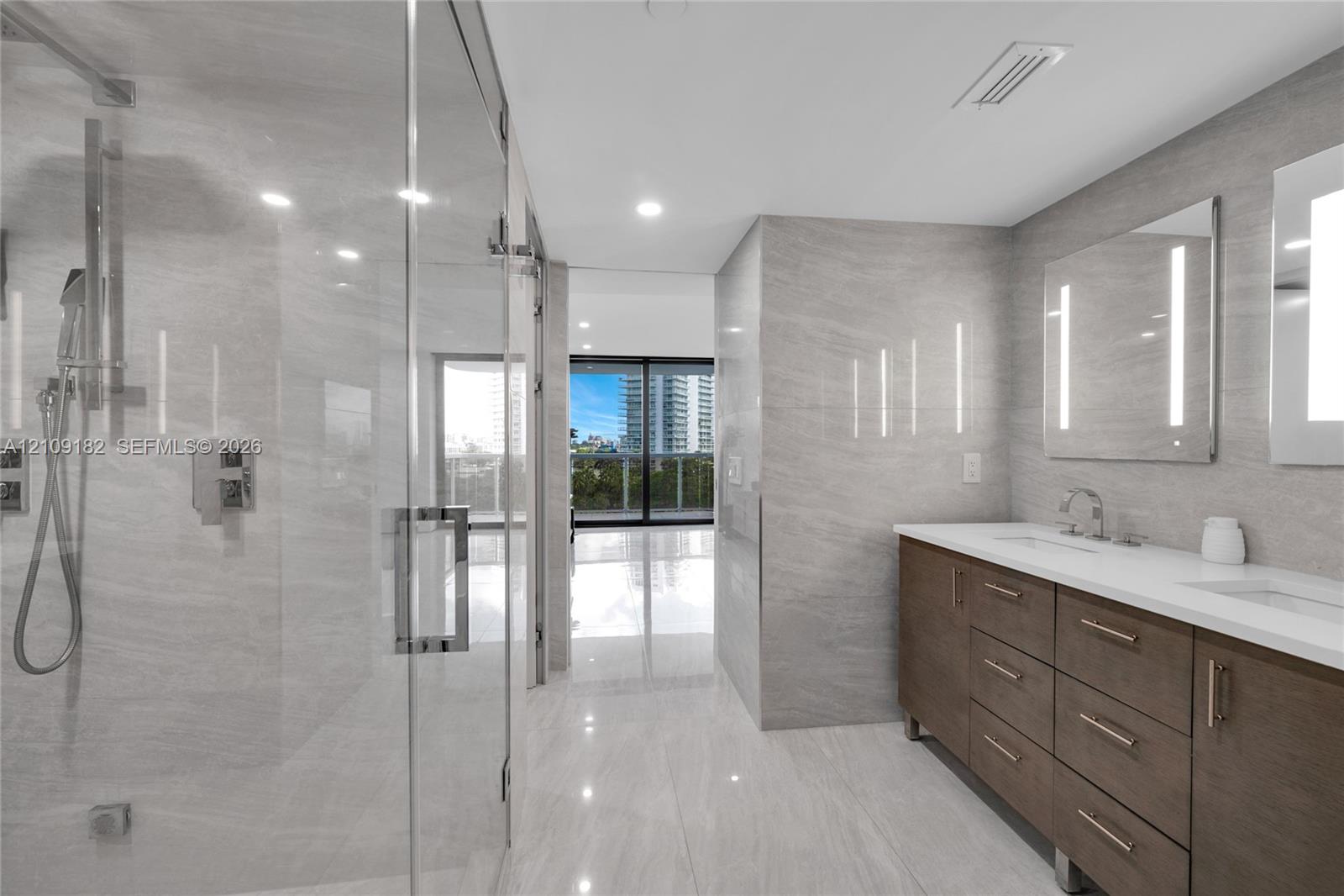 Double vanity with wood-finish cabinetry, white countertop, integrated dual sinks, and frameless glass-enclosed shower with tiled wall surfaces