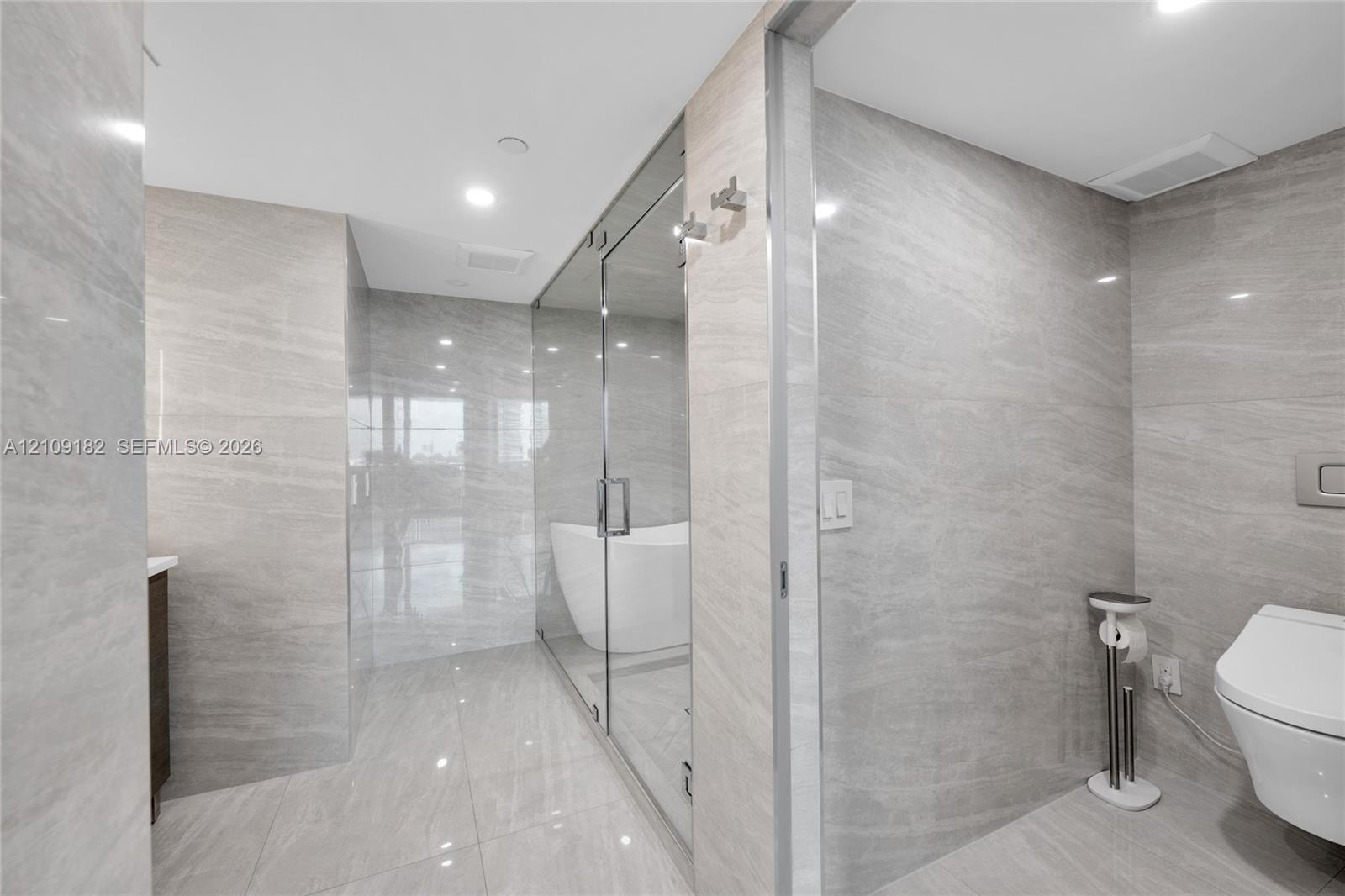 Bathroom featuring floor-to-ceiling large-format stone-finish tile, a glass-enclosed shower area, a freestanding soaking tub, and a wall-mounted toilet