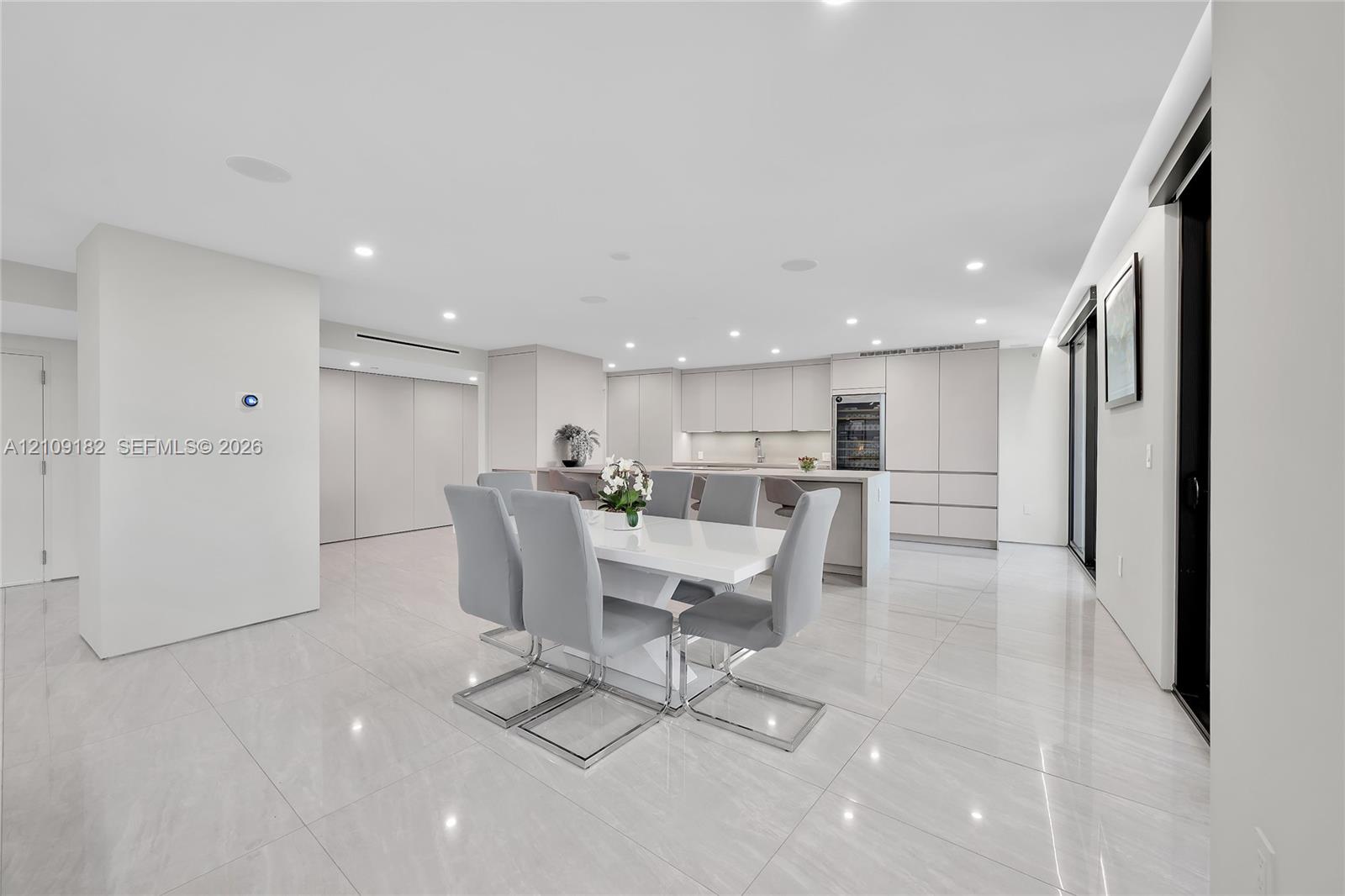Open-concept kitchen and dining area featuring large-format tile flooring, flat-panel cabinetry, a central island with integrated seating, recessed lighting, and a built-in wine refrigerator