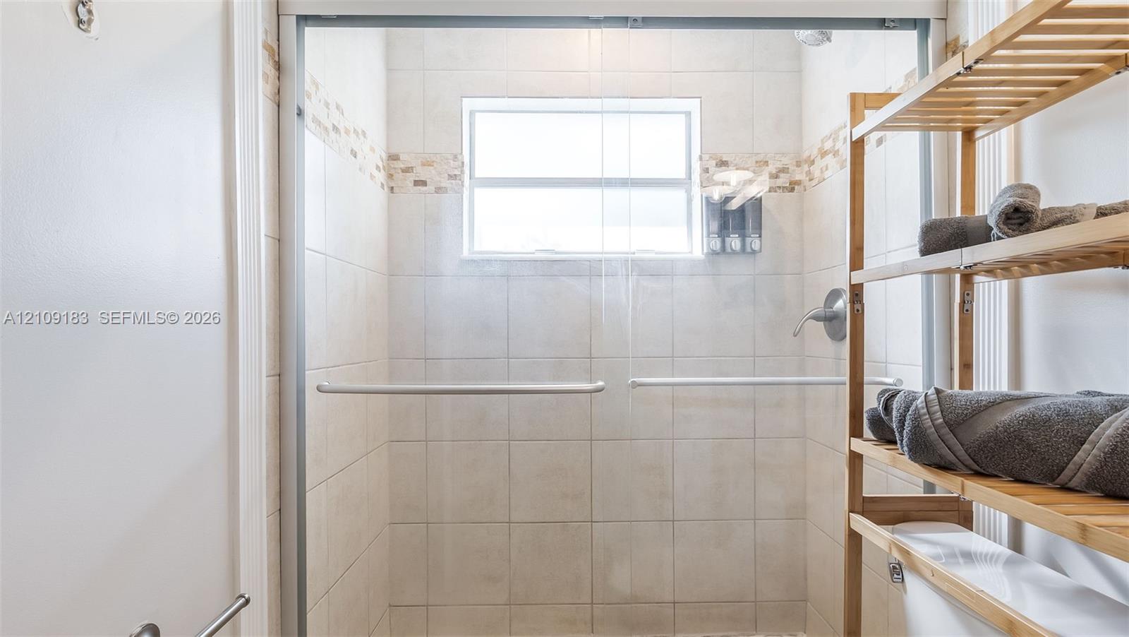 Walk-in shower enclosure featuring light-toned ceramic wall tile, horizontal mosaic tile accent banding, a framed glass door, and a built-in window