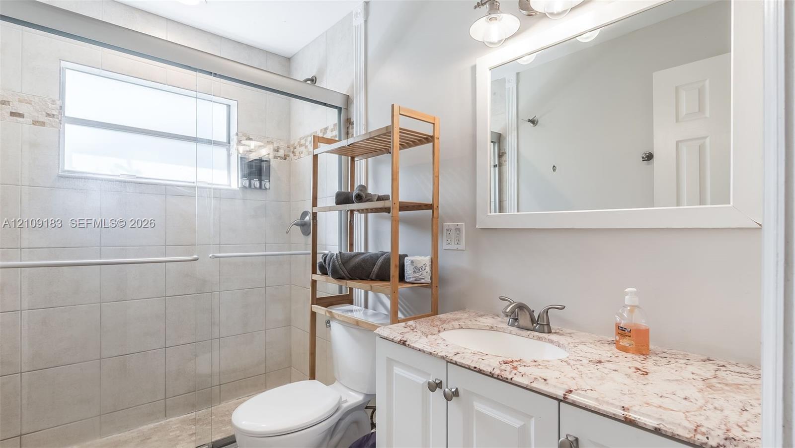Bathroom featuring a stone-surface vanity with an integrated sink, white cabinetry, a glass-enclosed shower with tiled walls, and a wall-mounted vanity mirror with integrated lighting