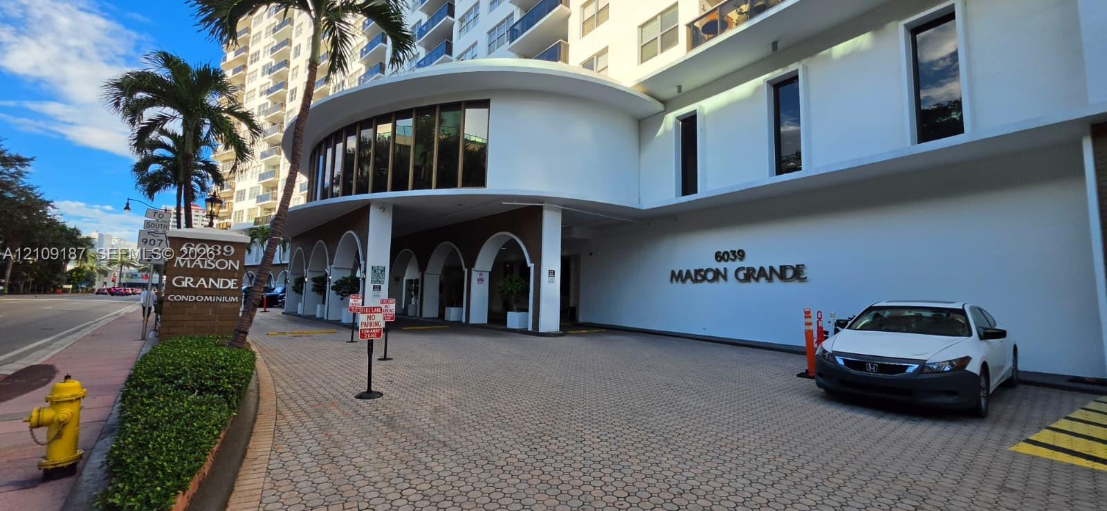 Exterior facade of the Maison Grande featuring a curved architectural silhouette, covered portico with arched supports, paved motor court, and multi-story balcony configurations