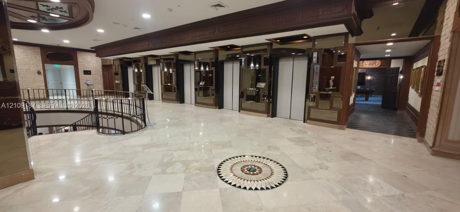 Grand lobby featuring polished stone tile flooring with decorative inlay, multi-elevator bank, ornate iron stair railing, and wood-paneled accents