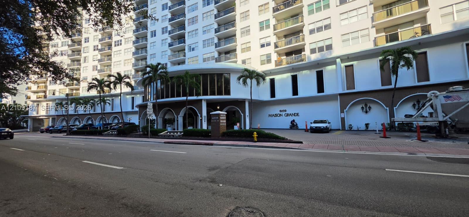Mid-rise residential building exterior featuring a porte-cochre entry, arched decorative wall niches, multi-story balconies, and lush palm tree landscaping