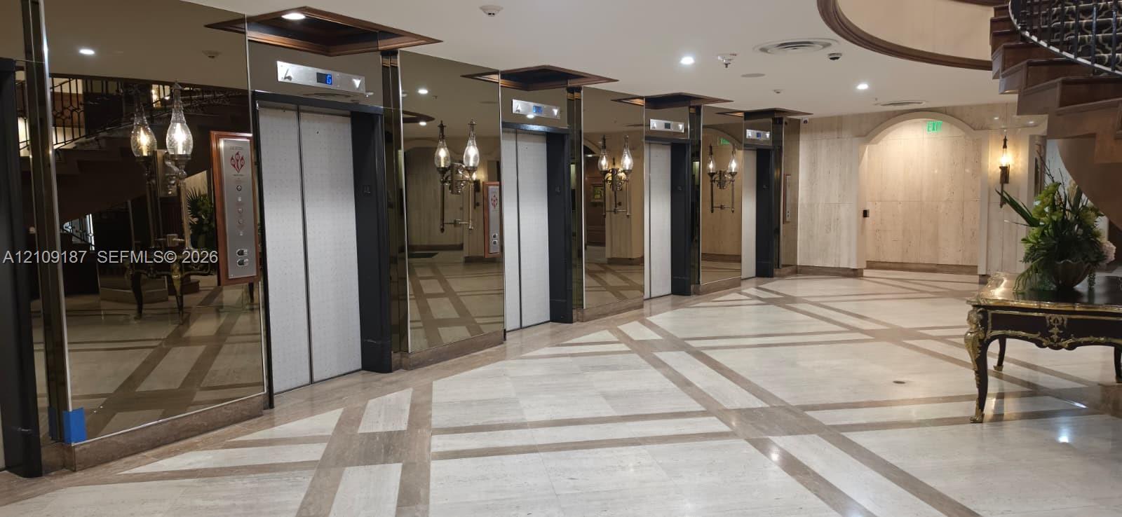 Grand lobby featuring floor-to-ceiling mirrored walls, a grid-patterned stone floor, a sweeping staircase, and multiple elevator banks