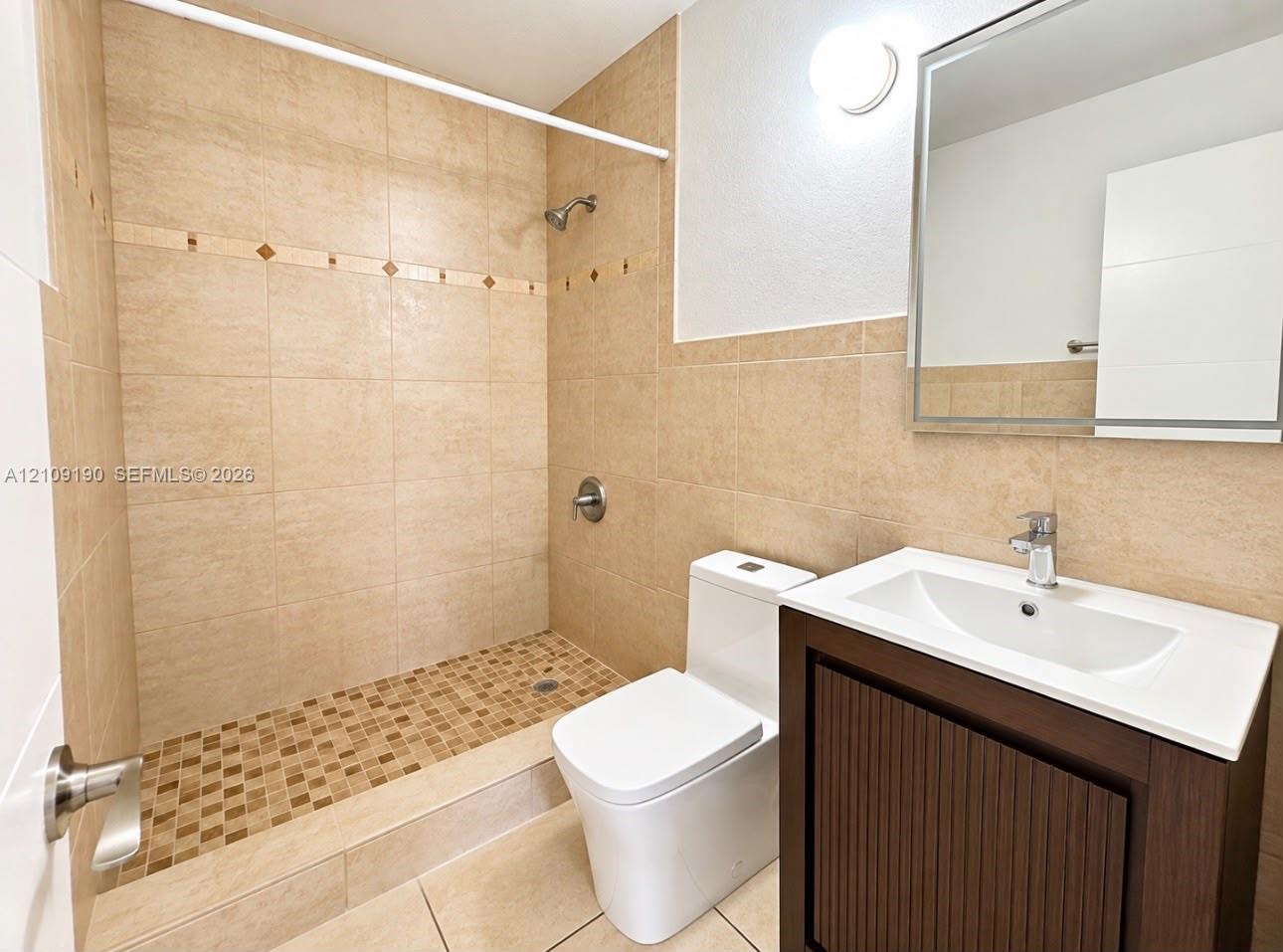 Walk-in shower with tiled surround and mosaic floor inlay, single-sink vanity featuring a solid surface countertop, and wall-mounted mirror