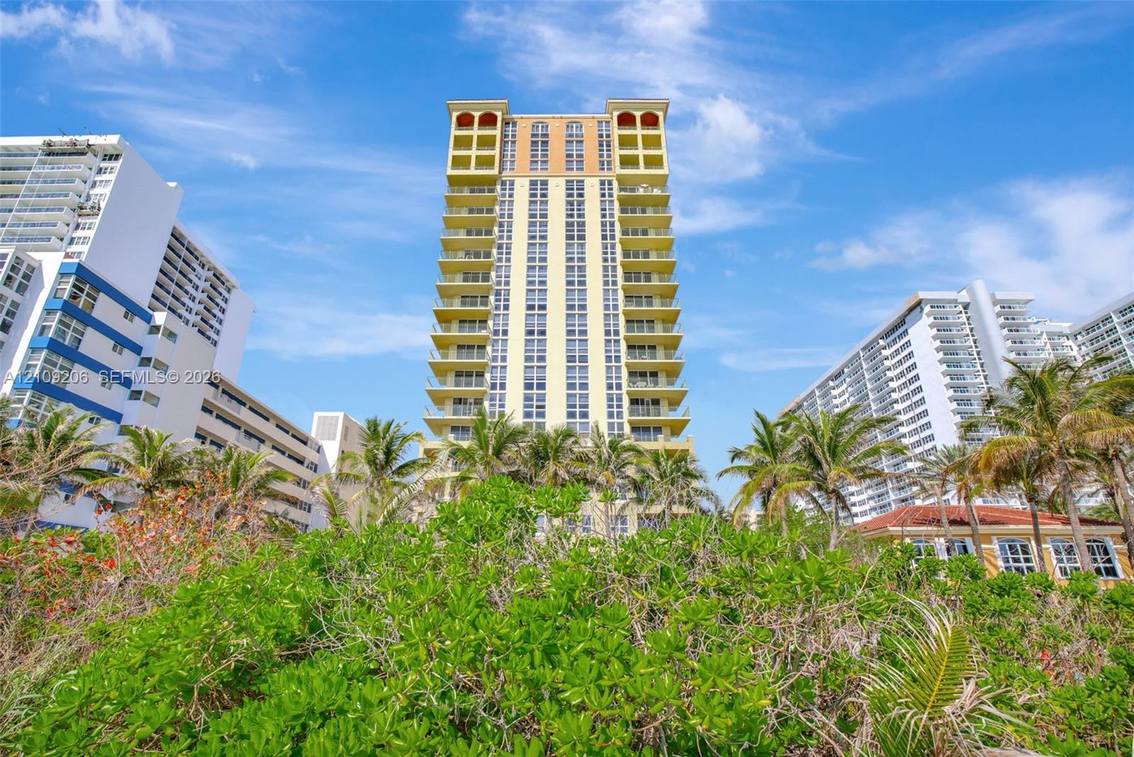 High-rise residential building featuring stacked private balconies, arched architectural detailing, and lush coastal landscaping
