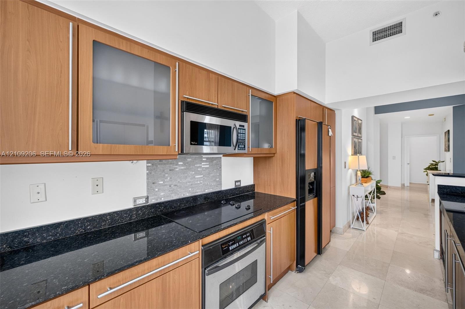 Kitchen featuring wood-finish cabinetry with frosted glass inserts, dark stone-finish countertops, tile backsplash, built-in stainless steel appliances, and tile flooring
