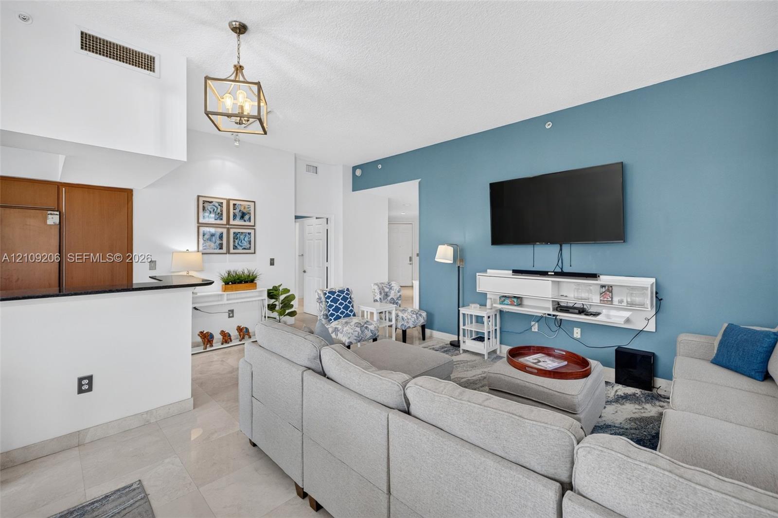 Living area featuring a high ceiling, metallic geometric pendant lighting, stone-finish tile flooring, a dual-tone accent wall, and a custom built-in white wall-mounted media console