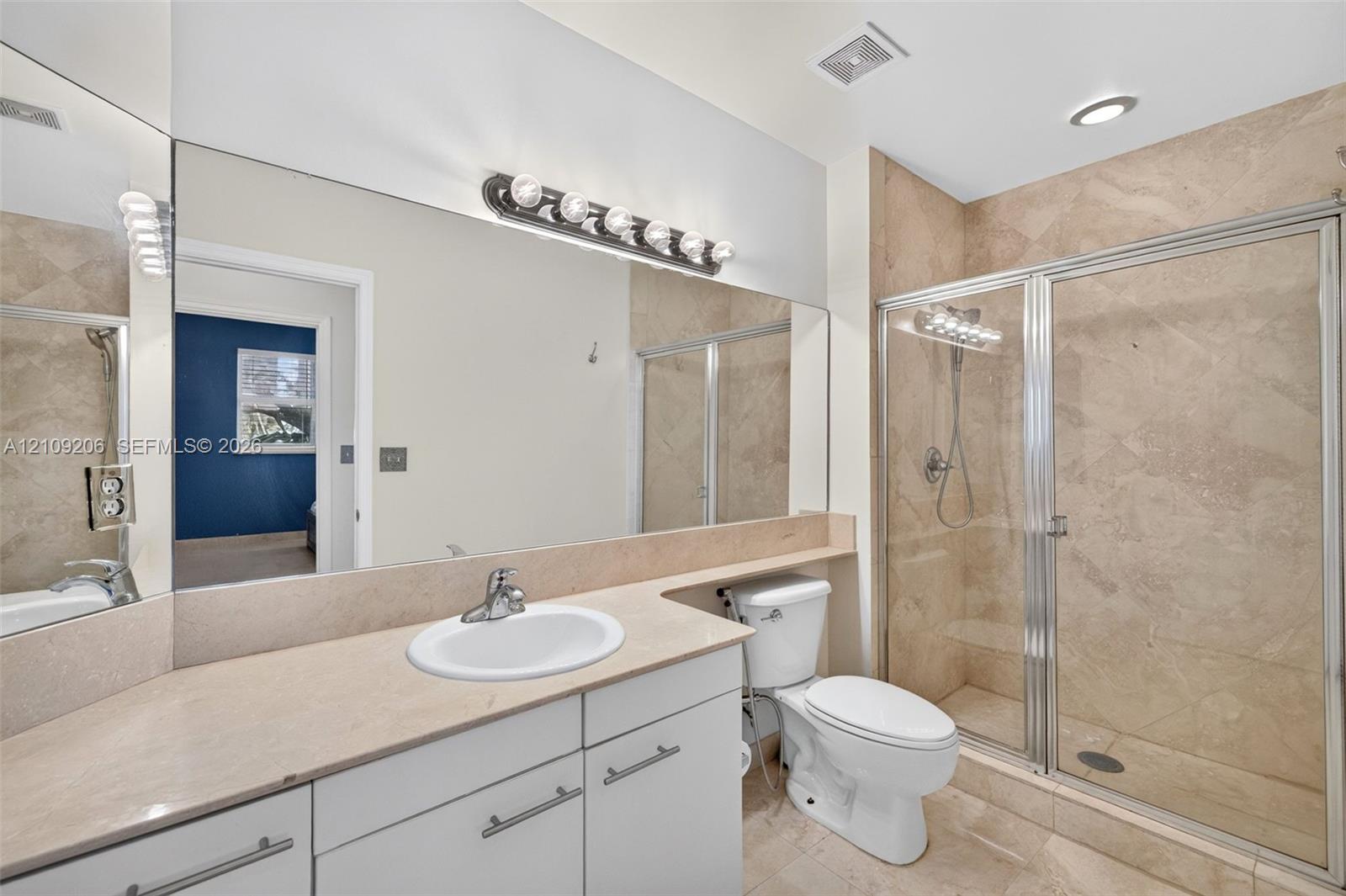 Full-width wall mirror above a vanity with stone-surfaced countertop, integrated sink, metal-finish hardware, and a walk-in shower enclosure featuring tiled walls