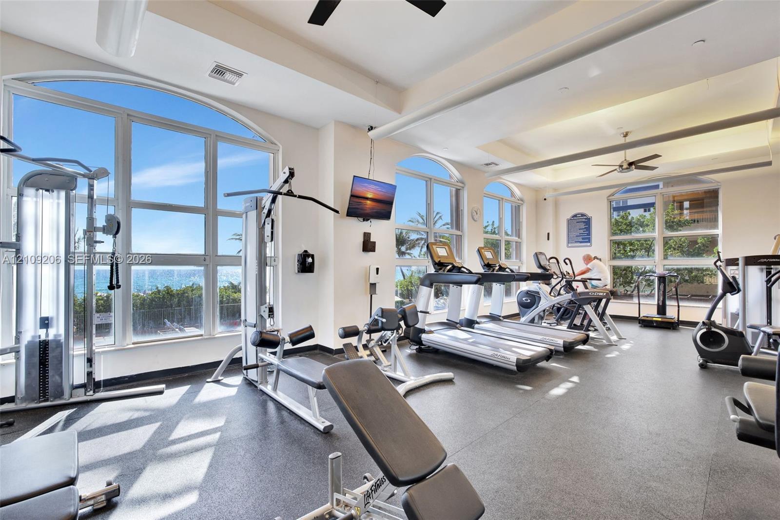 Fitness center featuring arched floor-to-ceiling windows, high tray ceilings, recessed lighting, and durable rubber flooring