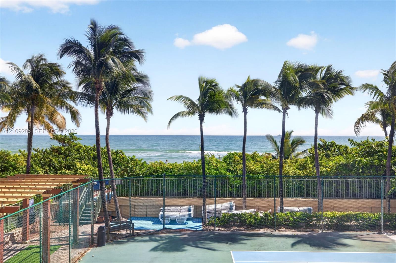 Tennis court enclosed by perimeter fencing, featuring a wooden pergola, mature palm trees, and ocean frontage