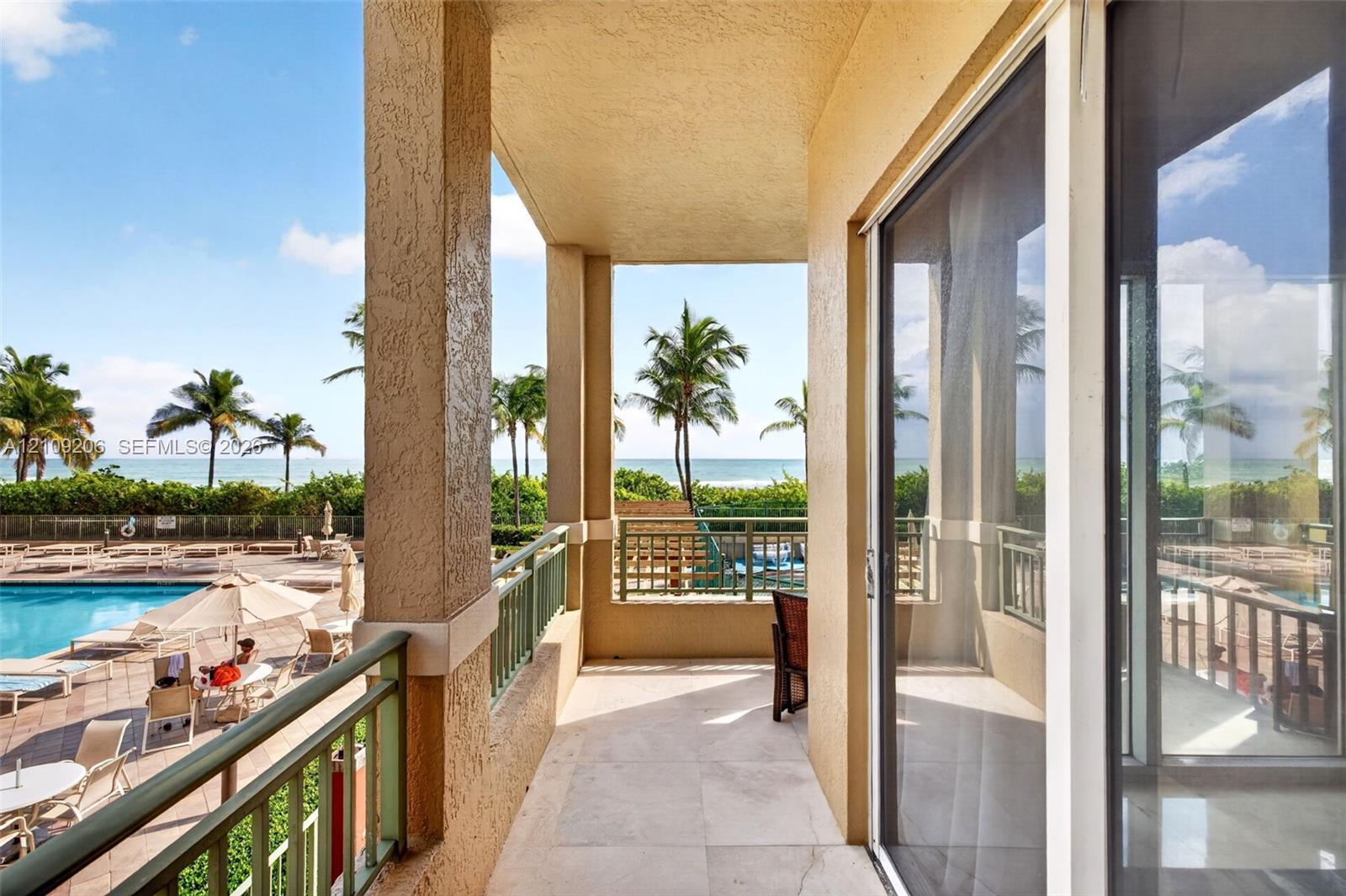Covered balcony featuring stone flooring and metal railings, overlooking an outdoor swimming pool and coastal scenery