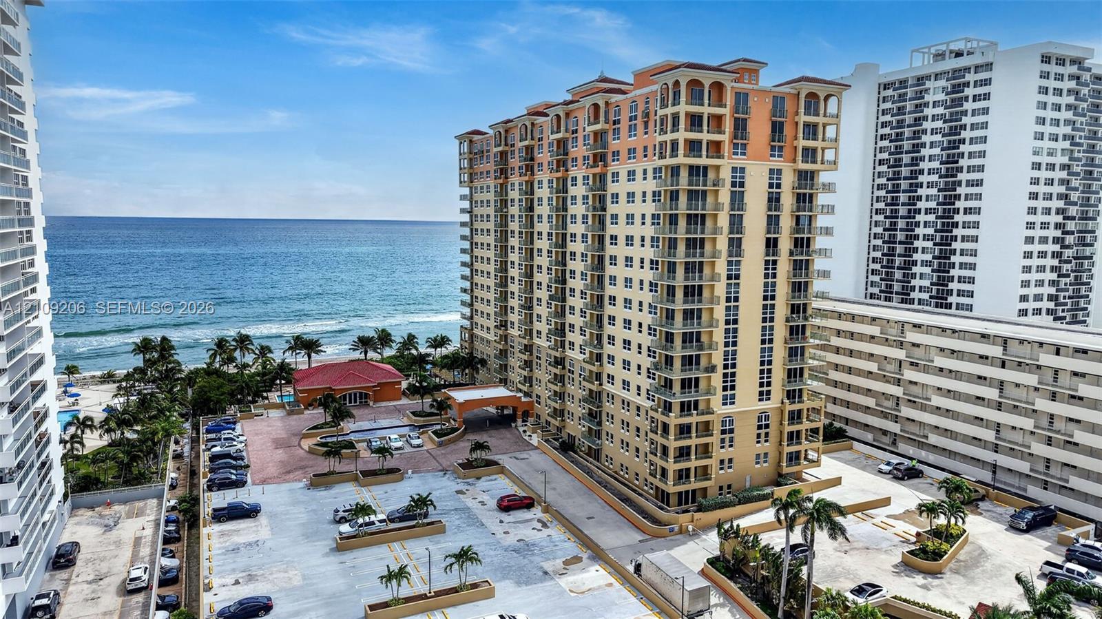 High-rise coastal building featuring private balconies, arched architectural detailing, a paved motor court, and proximity to the shoreline