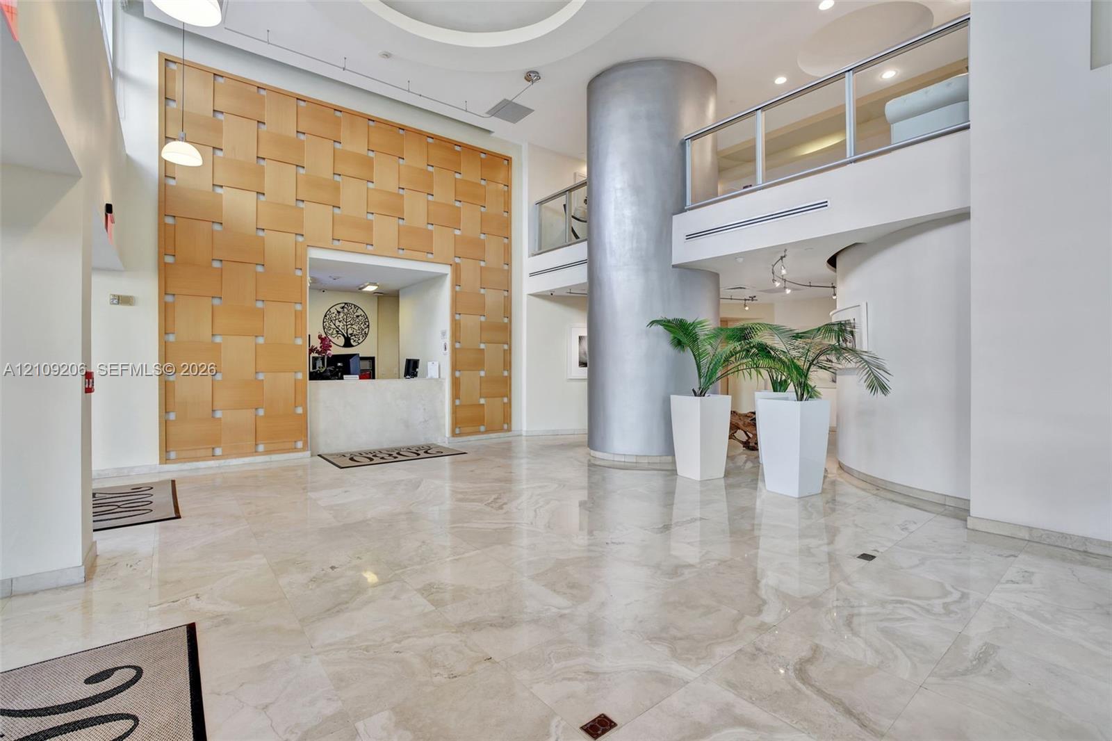 Grand entrance featuring polished stone flooring, a woven-wood accent wall, a cylindrical architectural column, and a mezzanine with glass-panel railings