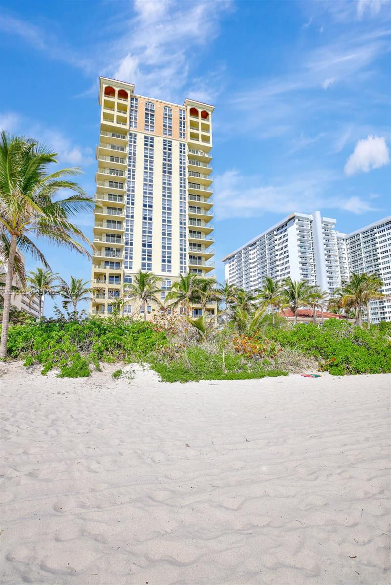 High-rise structure featuring stacked balconies, exterior arch details, sand-covered beach, and lush palm-lined vegetation
