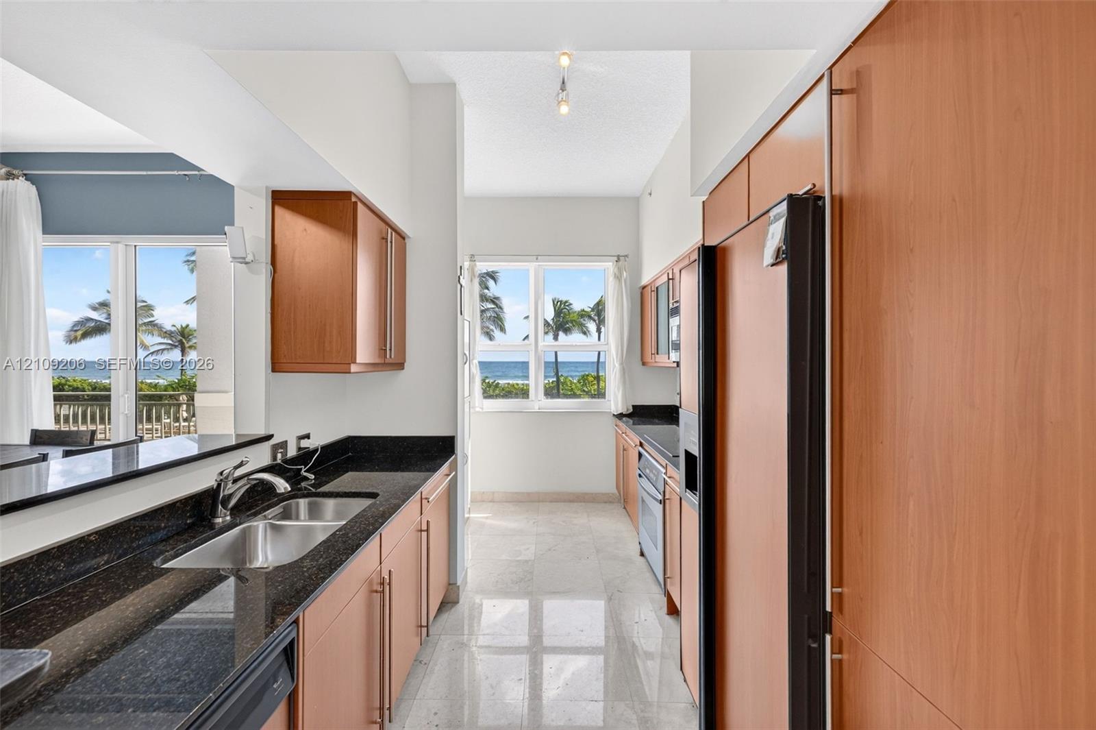 Kitchen featuring custom wood cabinetry, dark stone countertops, stainless steel double-basin sink, stone flooring, and windows with ocean scenery