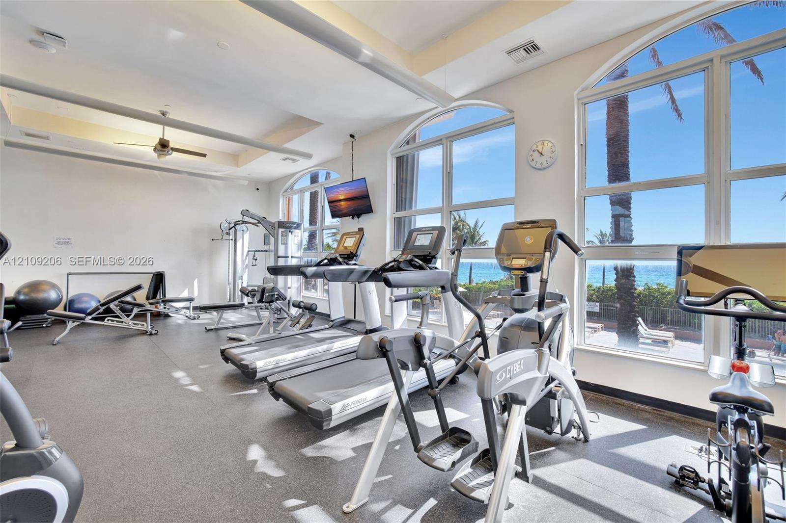 Fitness center featuring high ceilings, rubberized flooring, ceiling fan, and arched floor-to-ceiling windows with coastal scenery