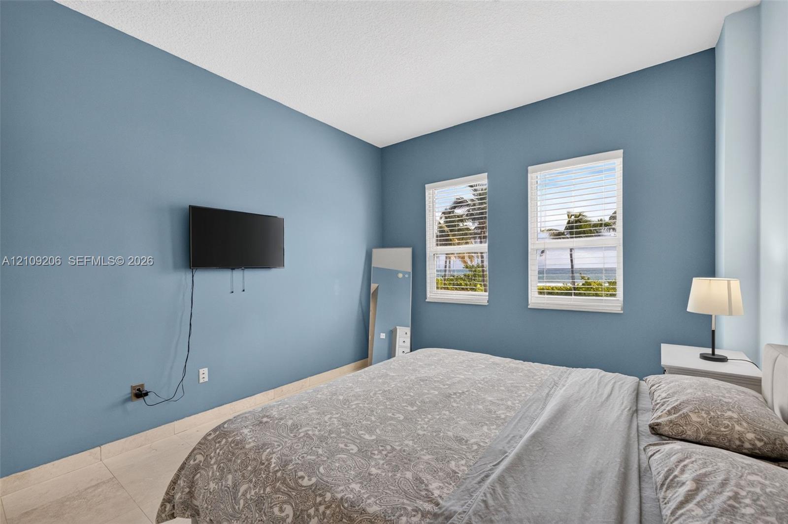 Spacious bedroom featuring light-toned tile flooring, two windows with horizontal blinds, and a coastal landscape visible through the glass
