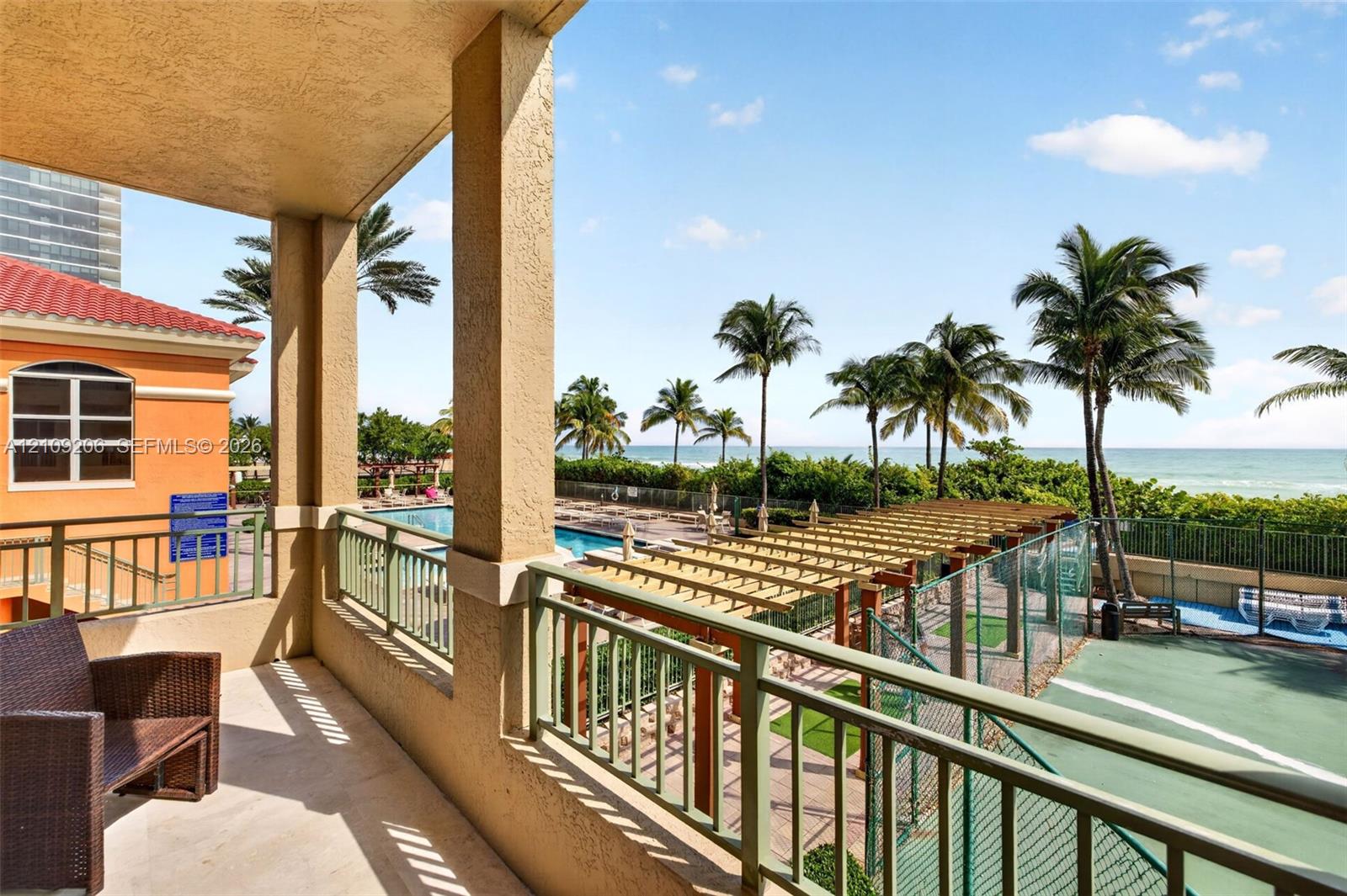 Covered balcony featuring metal railings, overlooking a tennis court, a wooden pergola, a swimming pool, and the ocean