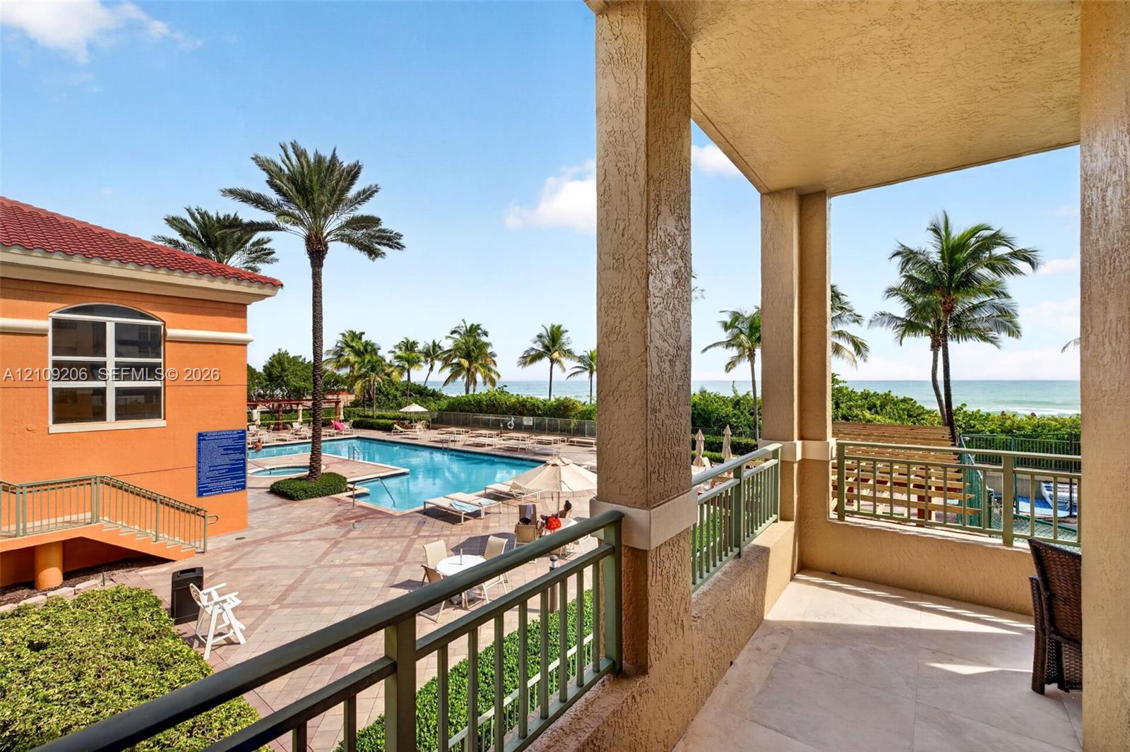 Covered balcony with stone flooring, metal railings, and stucco columns overlooking a swimming pool, palm trees, and ocean horizon