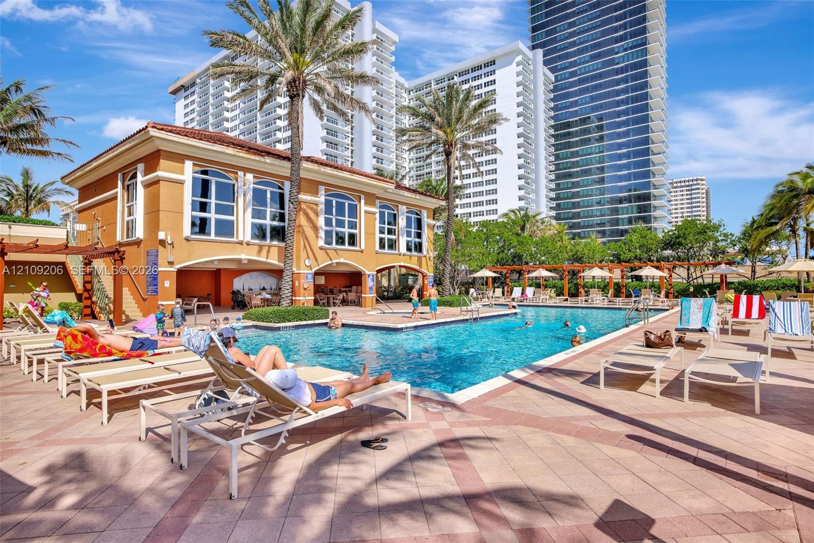 Swimming pool area featuring a stone-paver deck, shaded pergola structures, mature palm trees, and an exterior clubhouse building with arched windows