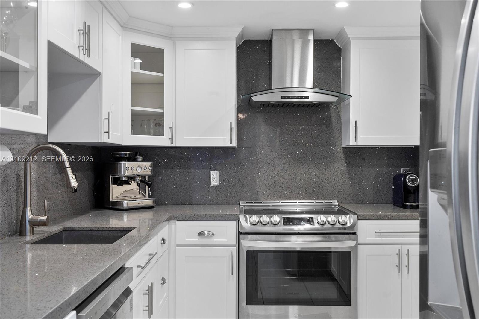 Kitchen featuring white shaker-style cabinetry, dark stone-finish countertops, full-height matching backsplash, stainless steel range with overhead chimney hood, and undermount sink with pull-down faucet