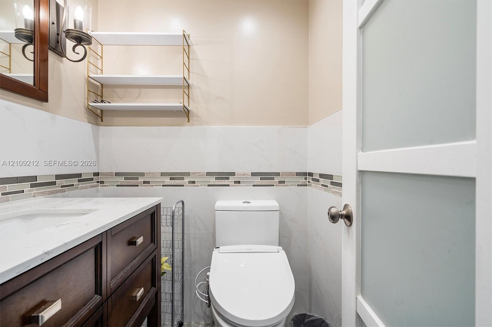 Dark wood vanity with a light-toned solid surface countertop, glass-paneled interior door, wall-mounted metal shelving, and tile wainscoting with decorative mosaic accent inlay